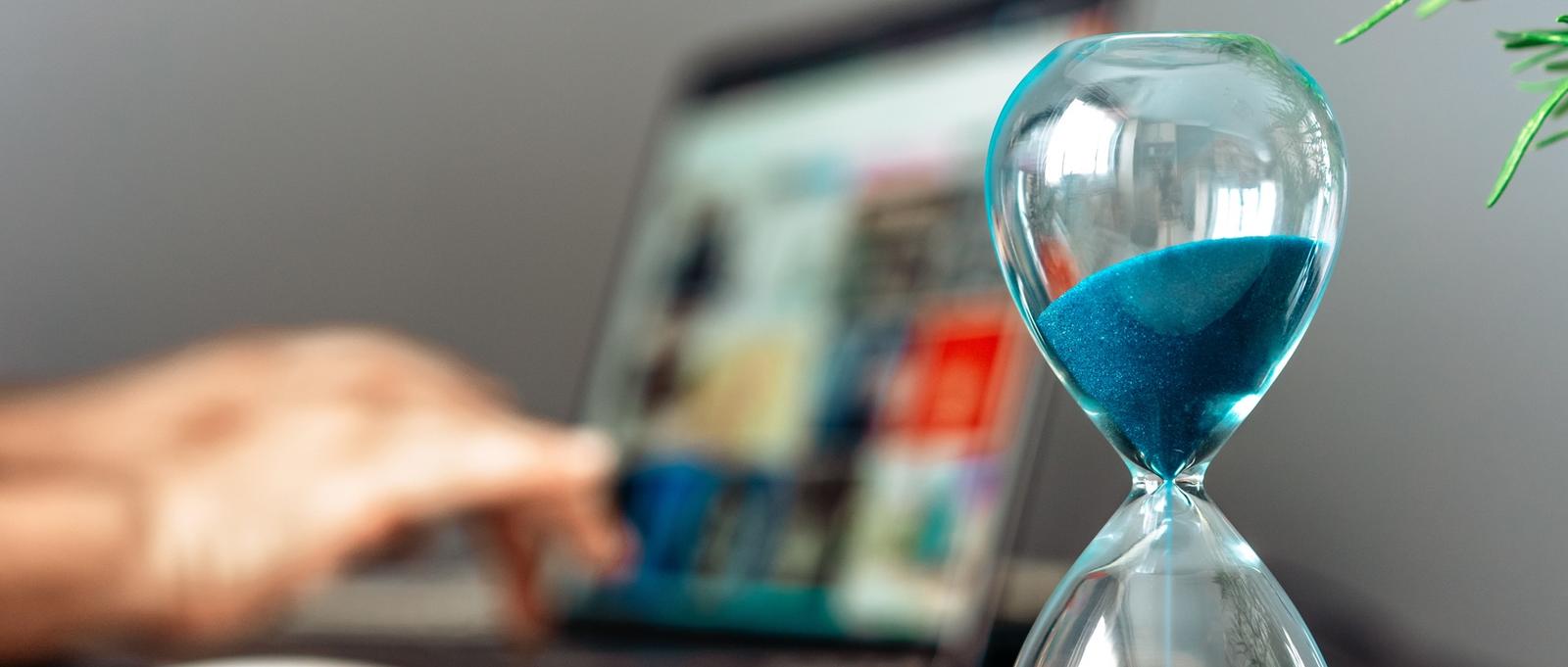 Sablier en verre avec du sable bleu à côté d'un ordinateur portable en cours d'utilisation, symbolisant la gestion du temps dans le travail numérique.