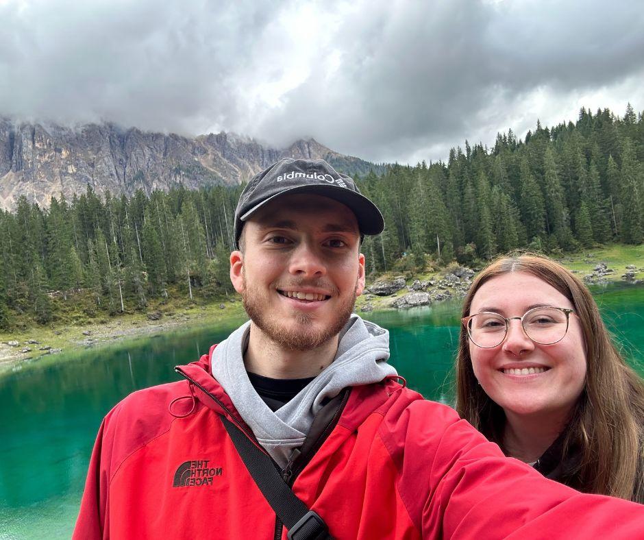 Jeune couple posant pour un selfie au bord d'un lac bordé d'arbres, au pied d'une chaîne de montagnes.