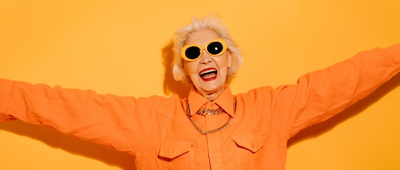 Une femme souriante, âgée et élégante, portant des lunettes de soleil à la mode, écarte les mains, veut serrer quelqu'un dans ses bras, heureuse de vous voir, isolée sur un fond jaune.