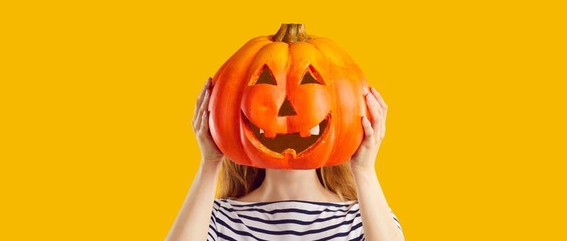 Femme en haut rayé sur fond jaune cachant son visage derrière une lanterne de citrouille d'Halloween.