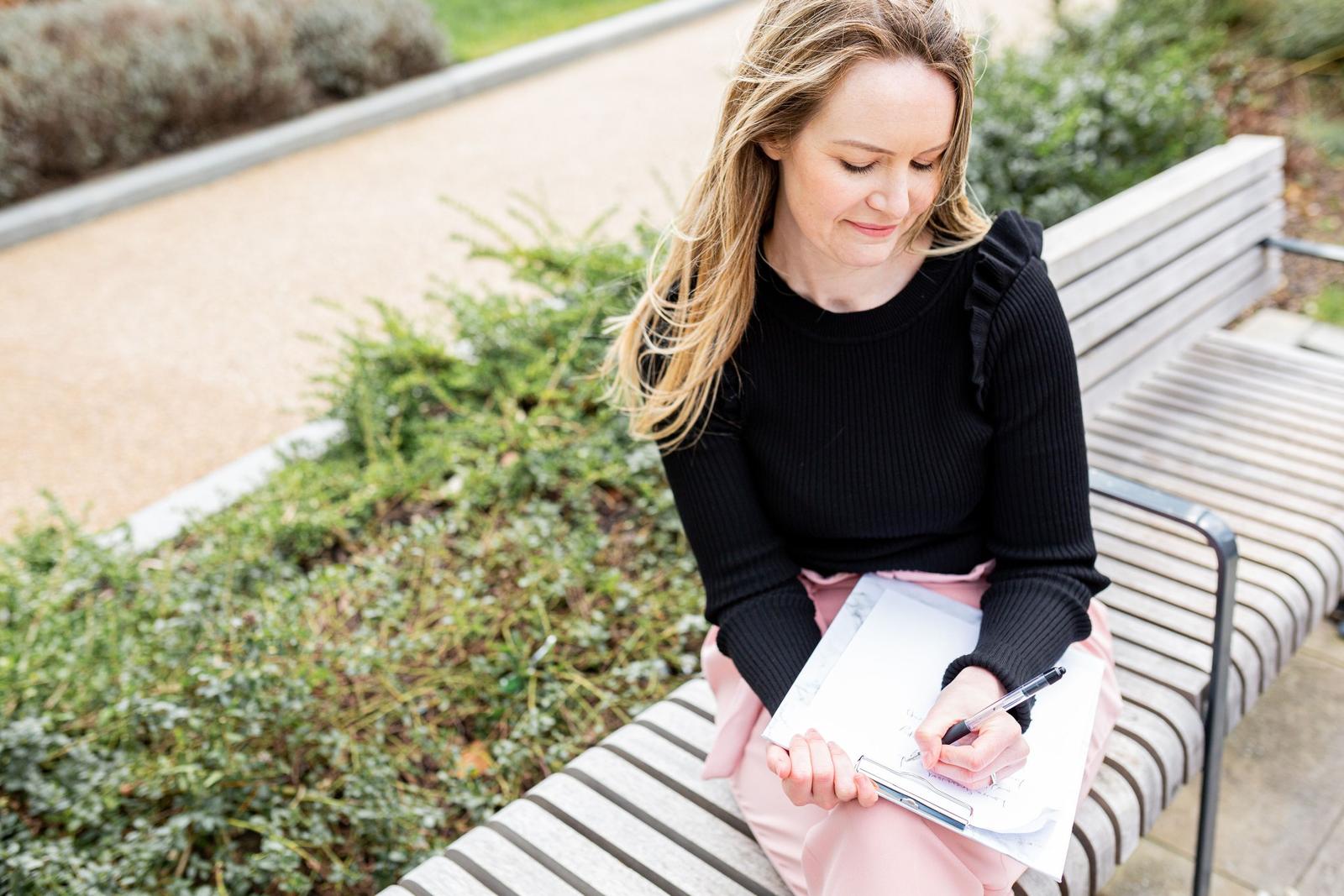 Psychothérapeute accréditée, Laura Greenwood de Laura Greenwood Therapy, assise sur un banc dans la nature avec un bloc-notes.