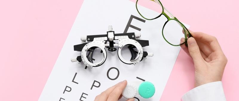Pair of ophthalmologist's hands holding eye test equipment - including eye chart, contact lens case, and green spectacles against a pink background.