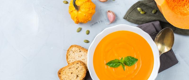 Soupe de potiron dans un bol avec des tranches de pain, un potiron, des graines de potiron, une cuillère en métal, une serviette bleue et des bulbes d'ail sur un fond gris/bleu pâle.