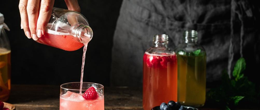 Versement à la main de kombucha à la framboise rose d'une bouteille dans un verre avec de la glace, avec des bouteilles de boissons colorées en arrière-plan.