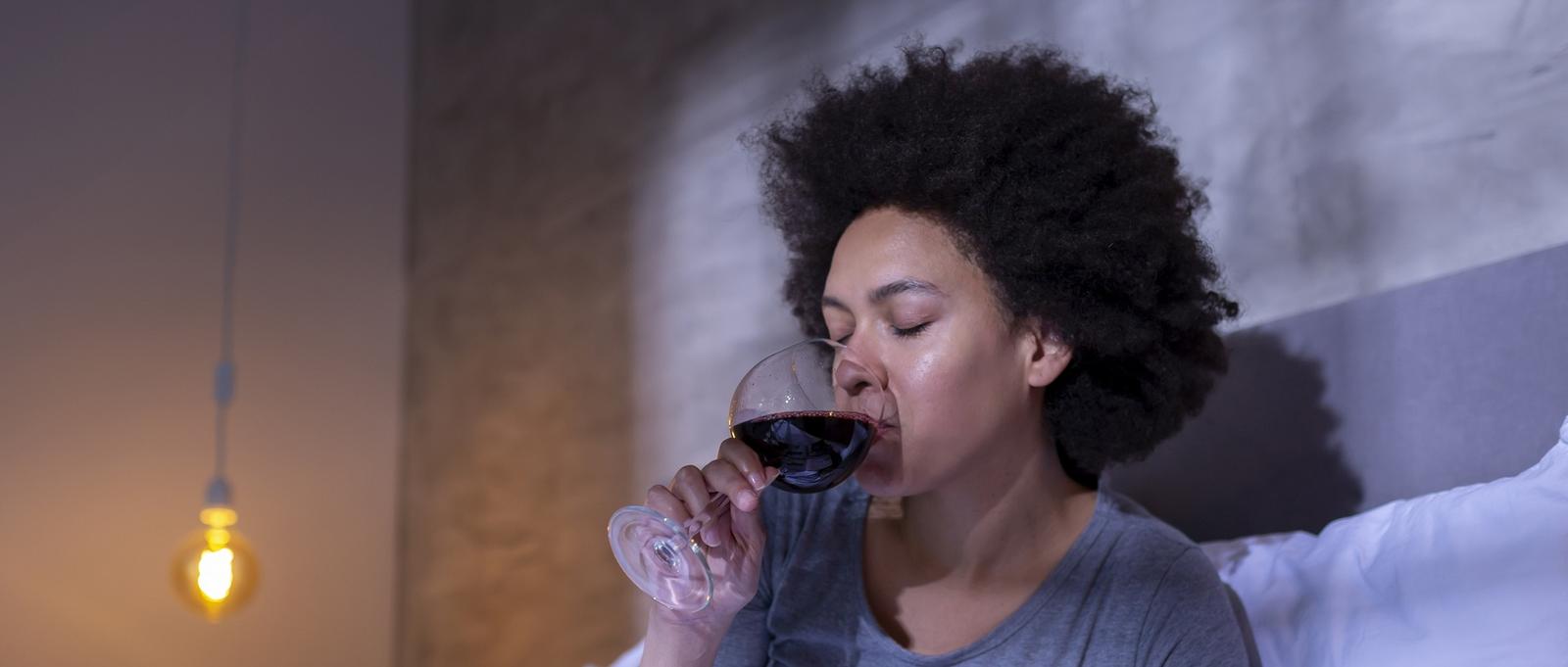 Personne aux cheveux bouclés sirotant un verre de vin rouge dans une pièce doucement éclairée par une chaude lumière suspendue.