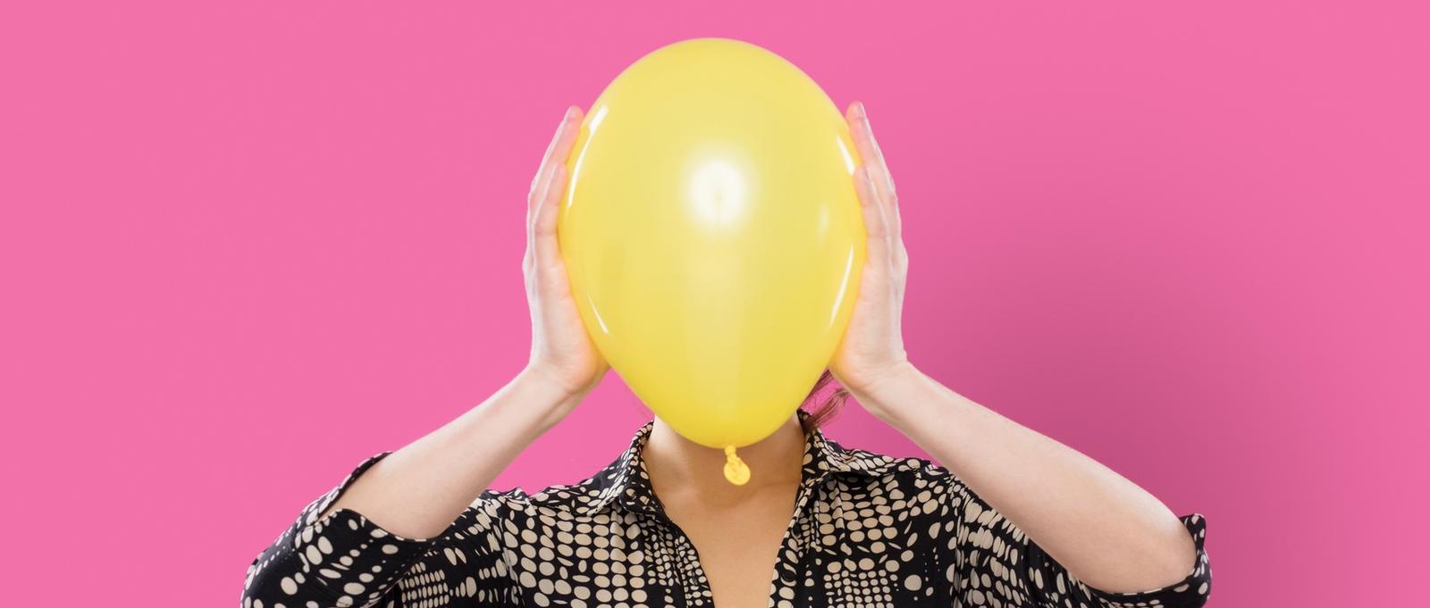 Femme sur fond rose tenant un ballon jaune sur le visage.