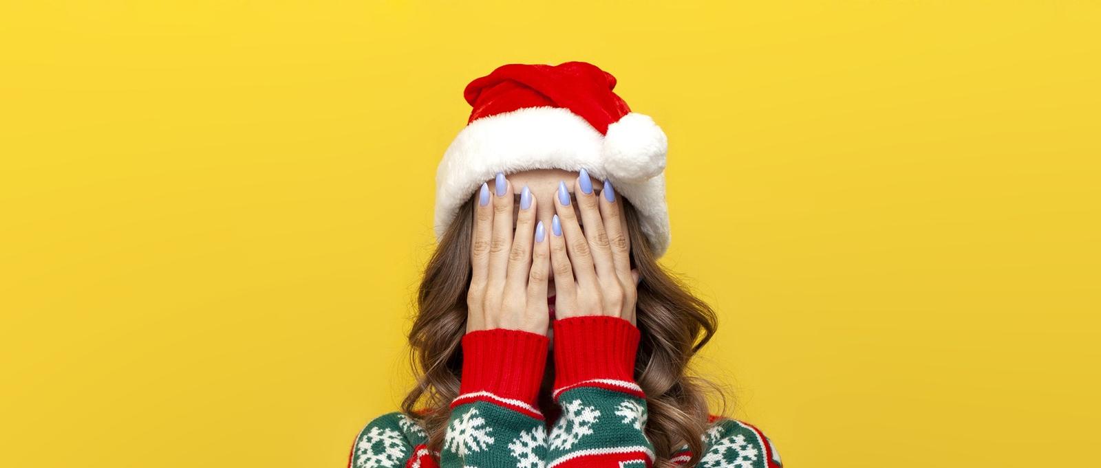 Une fille en pull de Noël et bonnet de Père Noël se couvre le visage avec sa main et est timide sur un fond jaune isolé, une femme se cache le visage en raison du stress et de la dépression dans ses vêtements du Nouvel An.