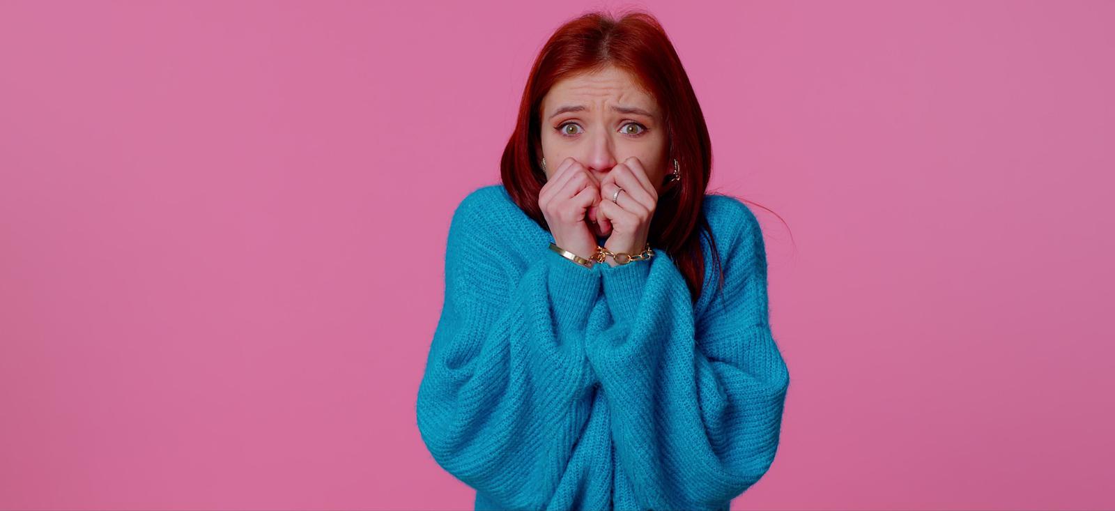 Person in bright blue jumper with worried expression covering mouth with hands against pink background.