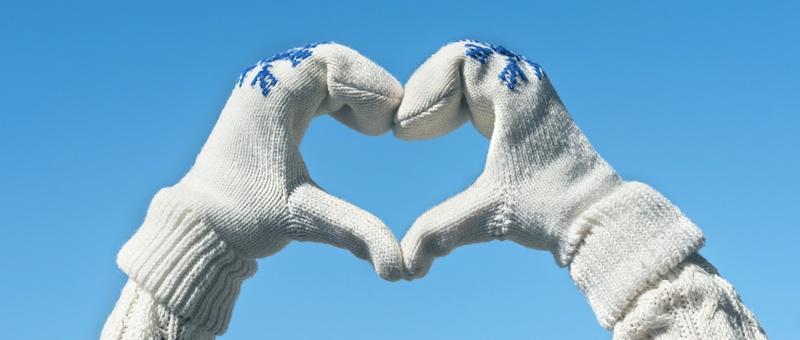 Personne faisant des mains en forme de cœur portant un pull blanc tricoté et des gants blancs tricotés avec des imprimés de flocons de neige, sur fond de ciel bleu hivernal.