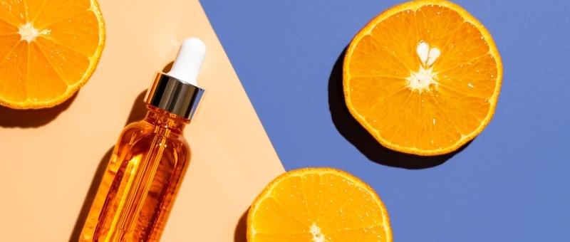 Flacon de sérum orange avec compte-gouttes à côté de moitiés d'orange fraîche sur fond pêche et bleu.