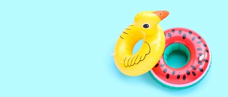Canard jaune gonflable et jouets de piscine en forme de pastèque sur fond bleu pâle.