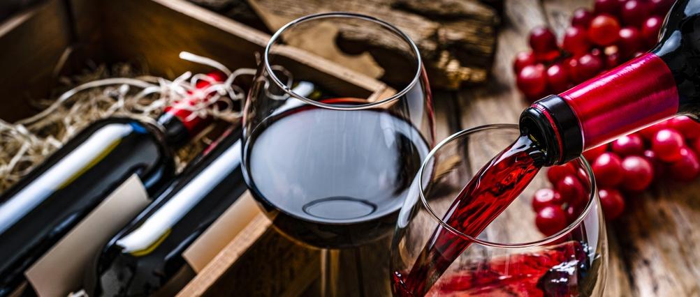 Vin rouge versé dans un verre à côté d'un verre de vin rouge rempli, avec des bouteilles et des raisins rouges frais sur une surface en bois.