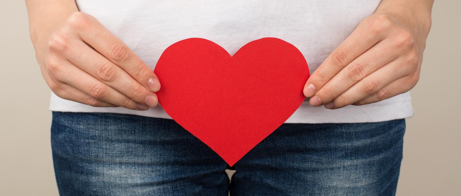 Photo en gros plan d'une femme en t-shirt blanc et jeans tenant un coeur en papier rouge près de l'entrejambe sur fond gris isolé.