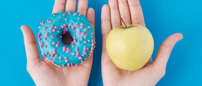 Pair of hands - one hand holding donut, one holding red apple against blue background.