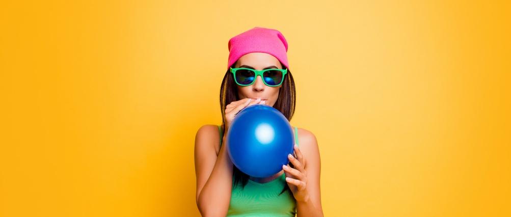 Happy funny hipster girl in pink hat inflate balloon near yellow wall