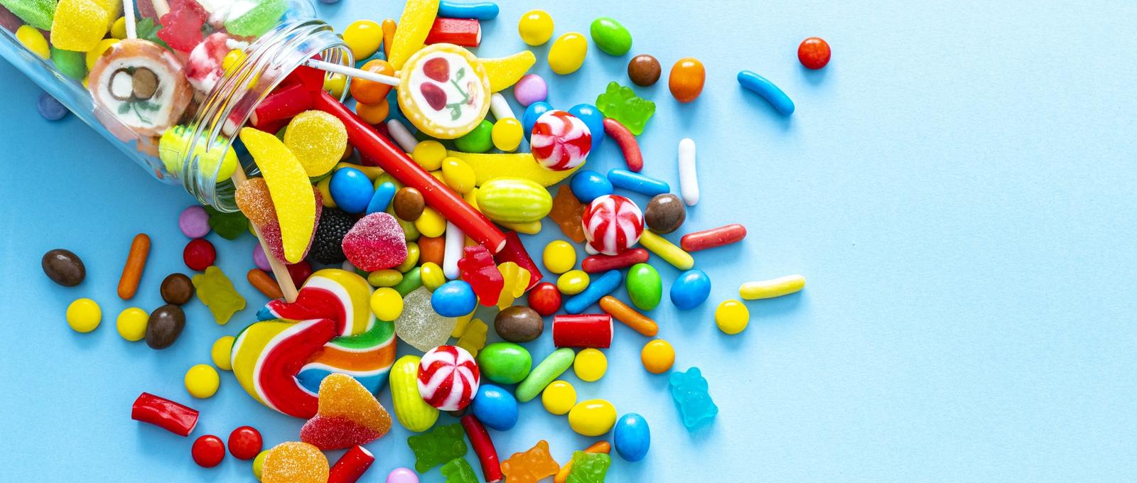 Grand groupe de bonbons multicolores s'écoulant d'un bocal en verre, filmés de haut en bas sur fond bleu.