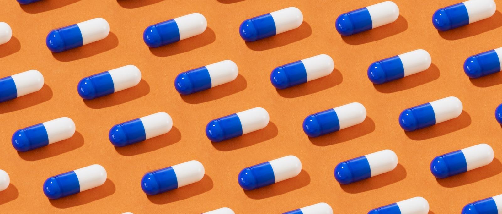 Des gélules bleues et blanches disposées en rangs bien ordonnés sur un fond orange, projetant de petites ombres.
