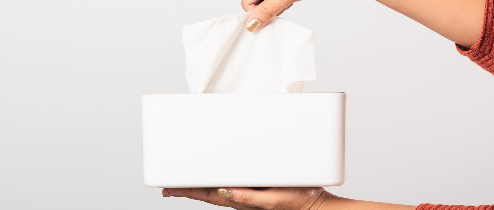 Main avec vernis à ongles doré tirant un mouchoir en papier d'une boîte rectangulaire blanche sur fond gris clair.