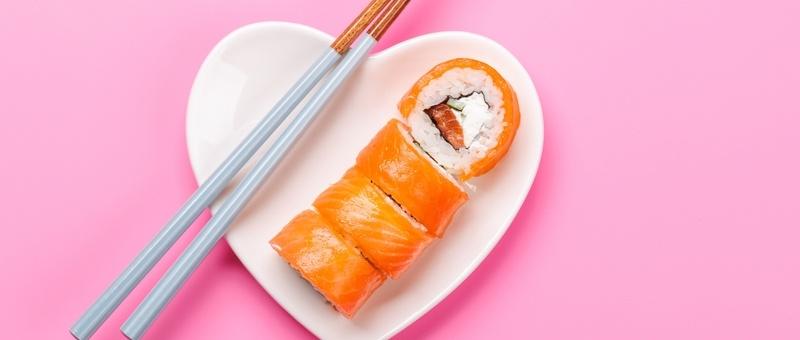 Salmon and cream cheese sushi with brown and grey/silver chopsticks on white heart-shaped plate against pink background.