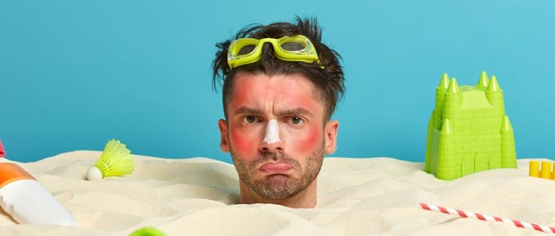 Homme enterré dans le sable avec des jouets de bord de mer et un visage brûlé par le soleil.