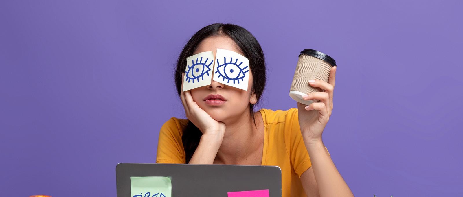 Portrait d'une jeune femme épuisée, assise sur son lieu de travail, avec des autocollants sur les yeux et une tasse de café à la main, travaillant sur un ordinateur portable à une table sur un fond de studio violet.