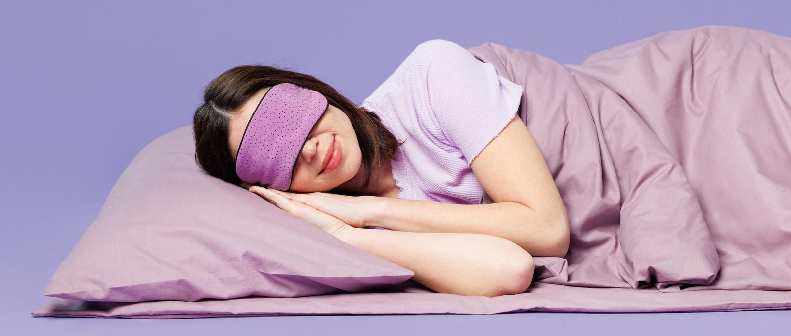 A female sleeping on the floor wrapped in a purple duvet, with a purple pillow and a purple eye mask.