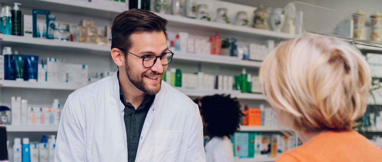 Jeune pharmacien discutant avec une cliente dans une pharmacie, avec une pharmacienne à l'arrière-plan. Étagères contenant des produits de santé.