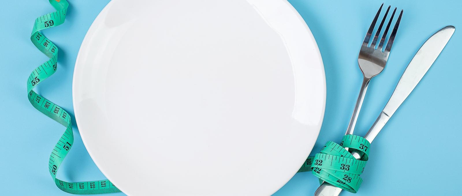 A empty white plate with a green tape measure next to it. On the other side of the plate is a fork and knife with the same tape measure wrapped around it. The image conceptualises eating disorders.