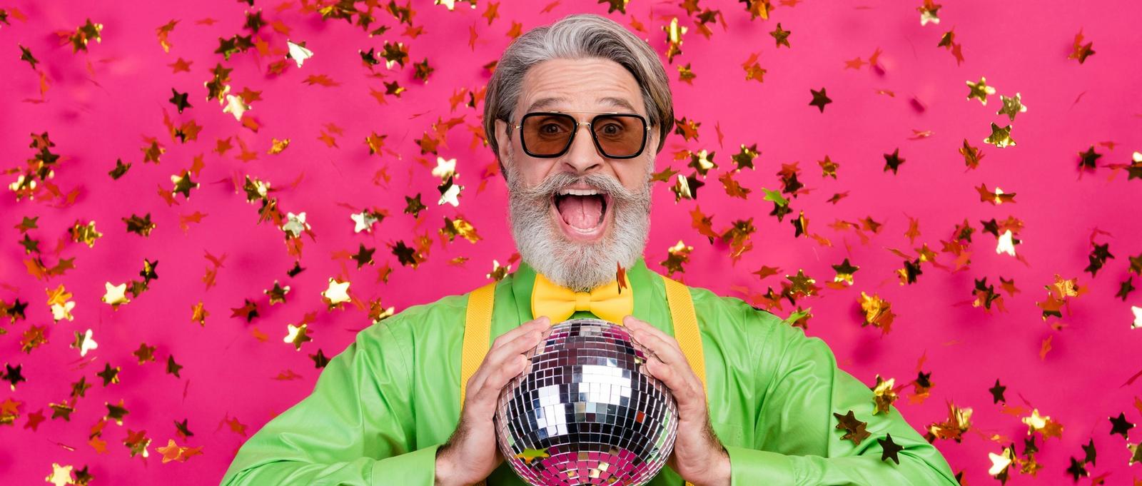 Photo d'un grand-père amusant en train de faire tomber des confettis, la bouche ouverte et excitée, tenant une boule disco lumineuse, portant des lunettes de soleil, une chemise verte, des bretelles jaunes et un nœud papillon, isolée sur un fond rose brillant.