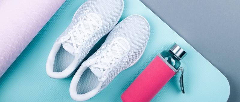 Vue de dessus de chaussures de sport blanches à côté d'une bouteille de boisson en métal rose reposant sur un tapis de yoga rose et bleu sur fond gris pâle.