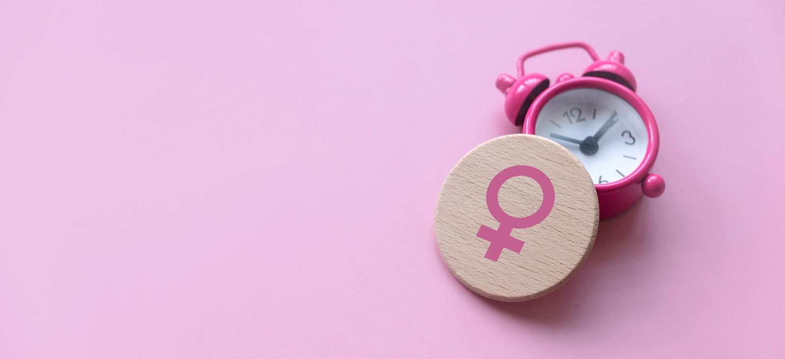 Pink alarm clock with wooden disc featuring female symbol on pink background, representing women's health timing.