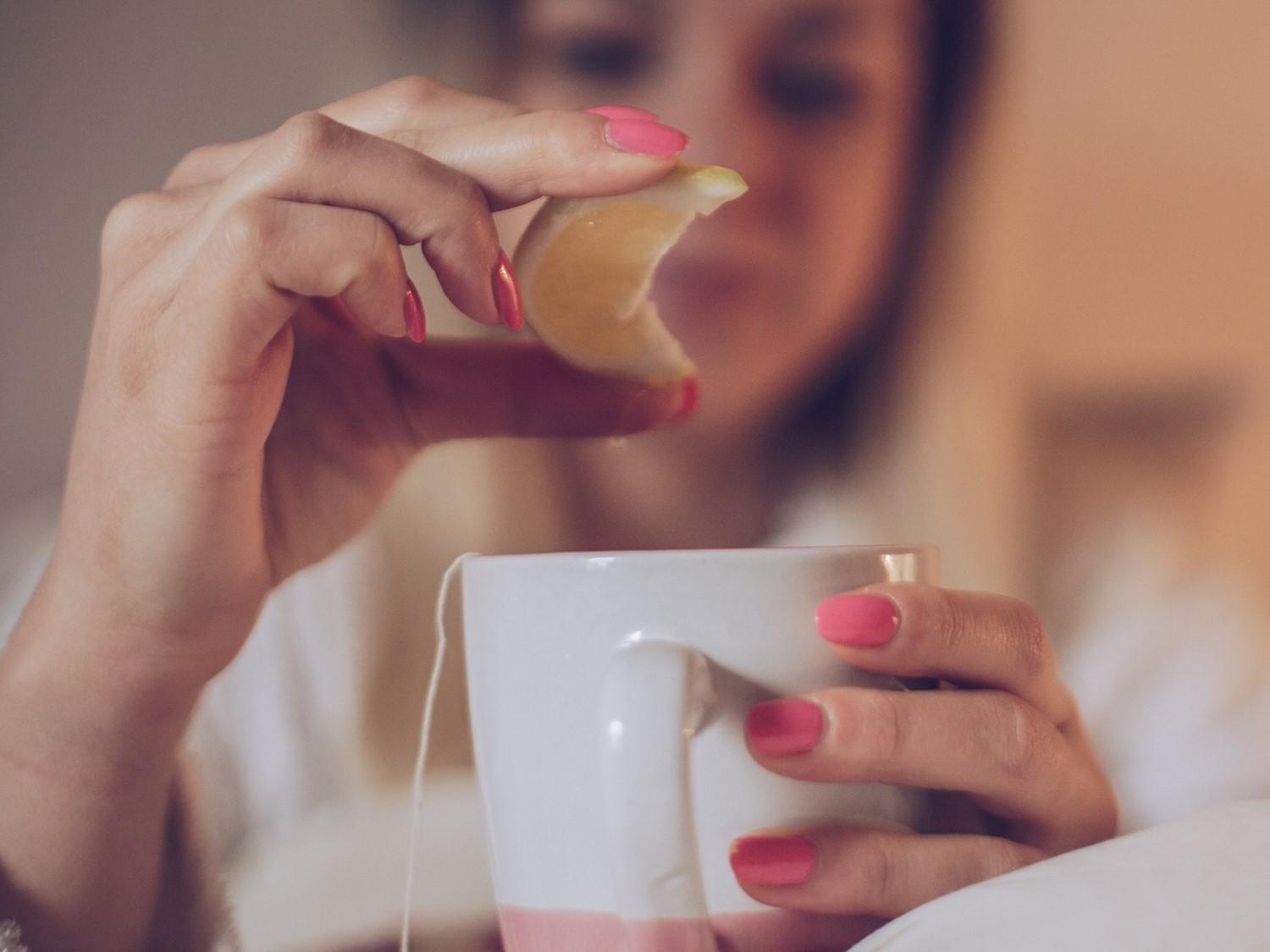 Personne aux ongles manucurés roses pressant du citron dans une tasse à thé blanche remplie de thé, arrière-plan flou.