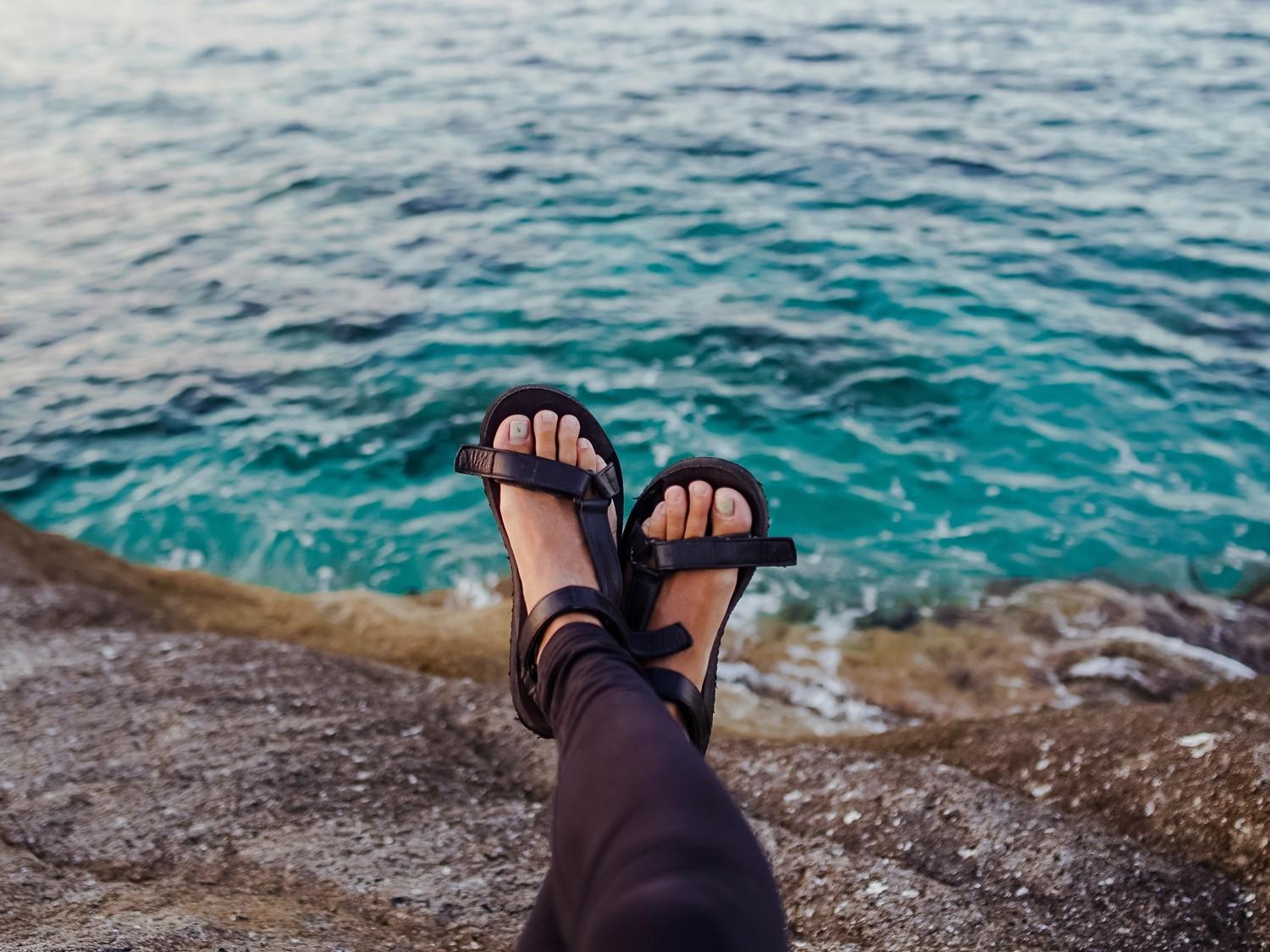 Pieds chaussés de sandales noires se balançant sur l'eau turquoise de l'océan, vue d'un bord de mer rocheux.