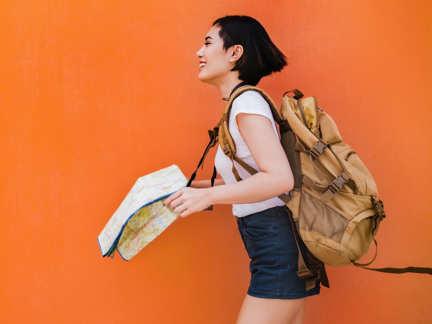 Personne aux cheveux bruns courts portant un sac à dos et tenant une carte, souriant sur un fond orange vif.