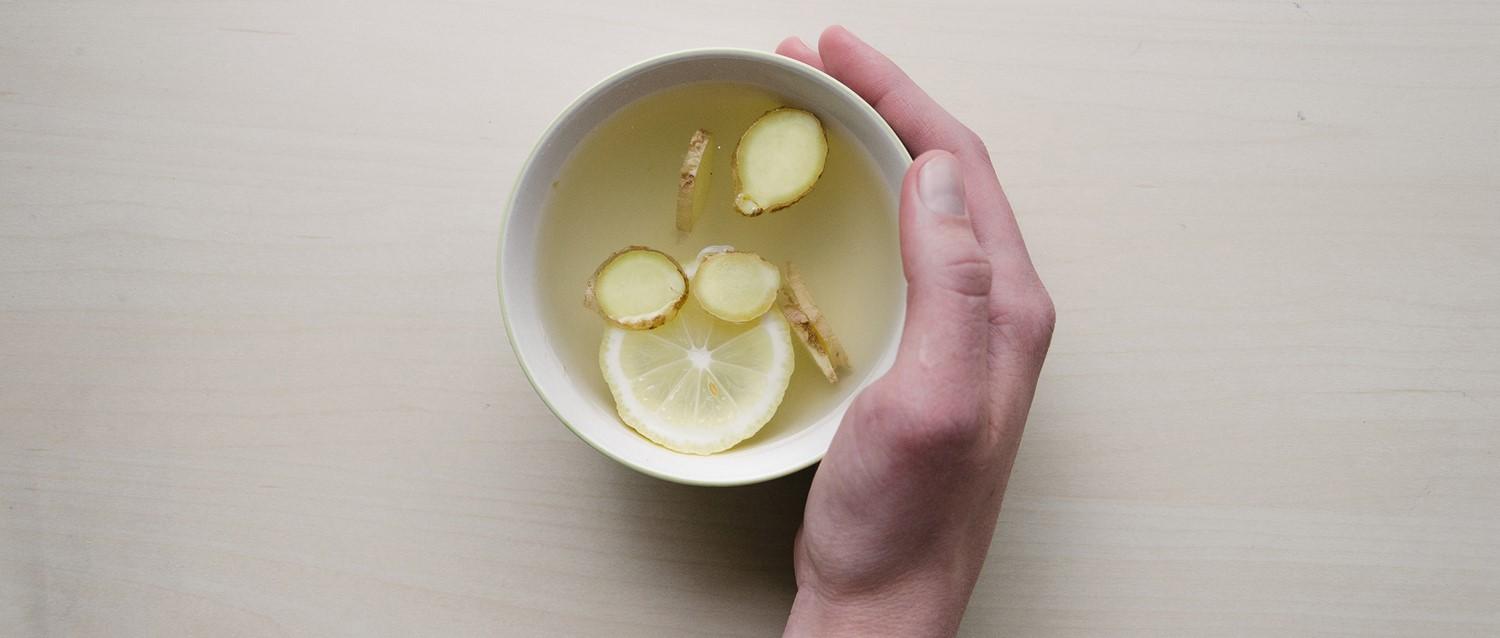 Main tenant un bol blanc de thé au gingembre et au citron avec des ingrédients tranchés flottant dans un liquide clair.