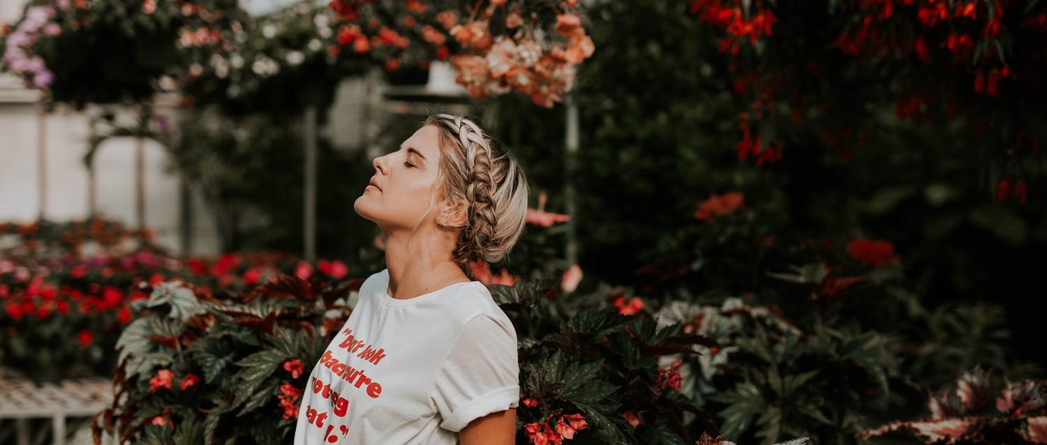 Femme aux cheveux blonds tressés, vêtue d'un tee-shirt blanc, entourée de fleurs d'un rouge éclatant et de verdure luxuriante dans un jardin.