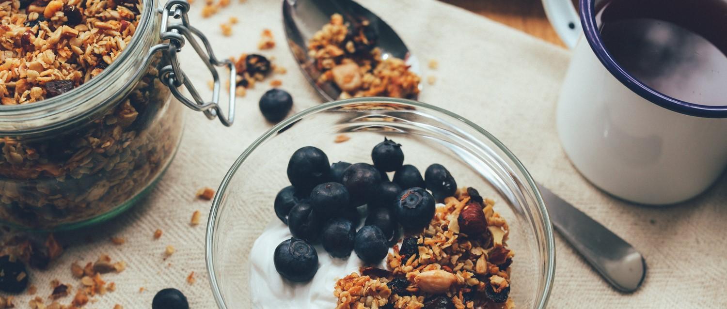 Des myrtilles fraîches et du granola maison dans un bol en verre avec du yaourt, ainsi qu'un pot de granola.