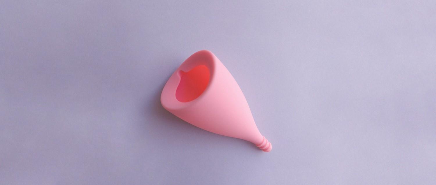 Coupe menstruelle rose posée sur un fond violet clair, montrant sa forme conique et son intérieur creux.