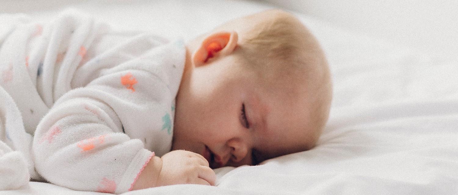 Nourrisson endormi en pyjama à motifs blancs, reposant paisiblement sur une literie blanche, avec une petite main recroquevillée près de son visage.