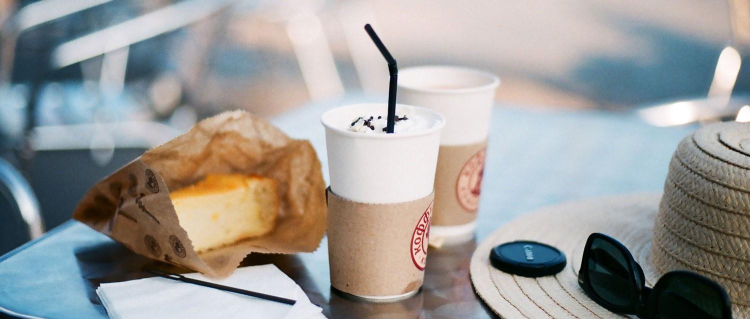 Tasses de café avec de la crème fouettée, pâtisseries dans un sac en papier, lunettes de soleil et chapeau de paille sur une table de café.