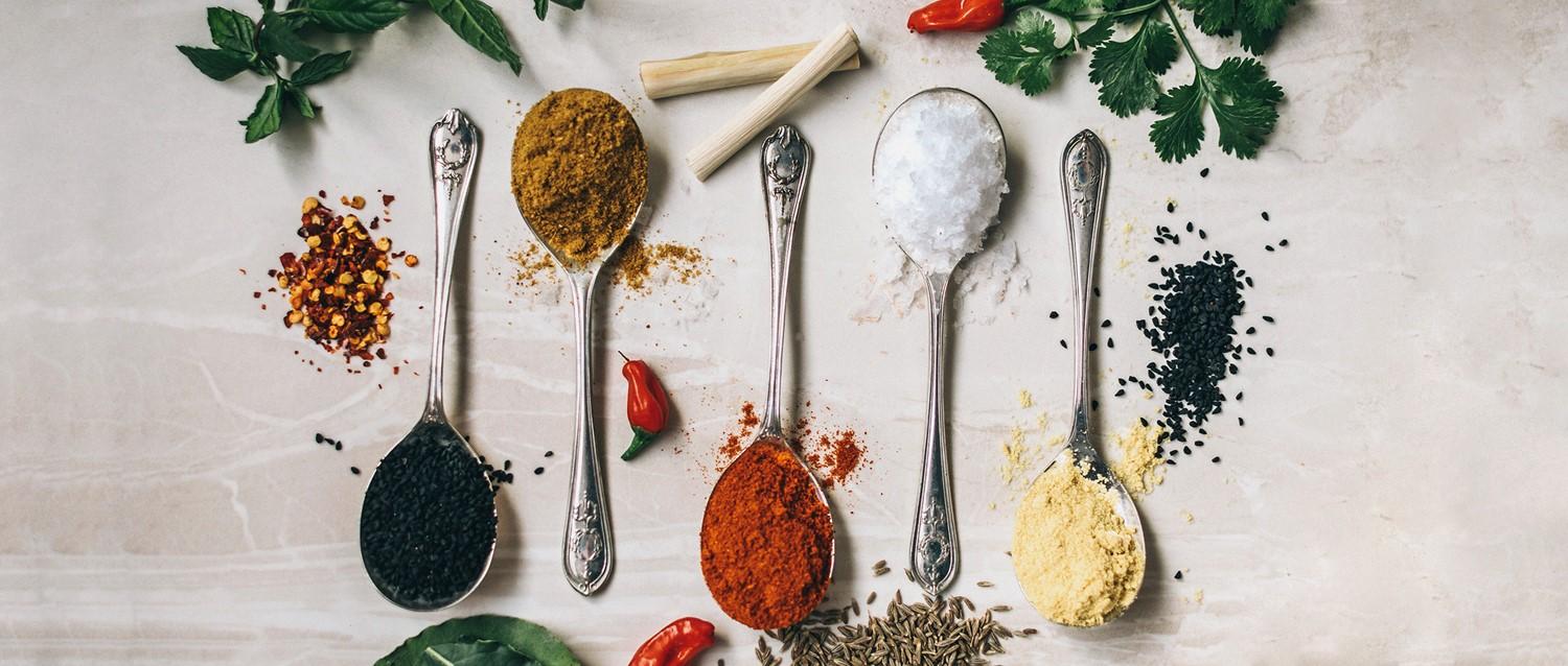 Cuillères en argent contenant des épices colorées, notamment du curry, du paprika, du sel et des graines noires, entourées d'herbes fraîches et de piments.