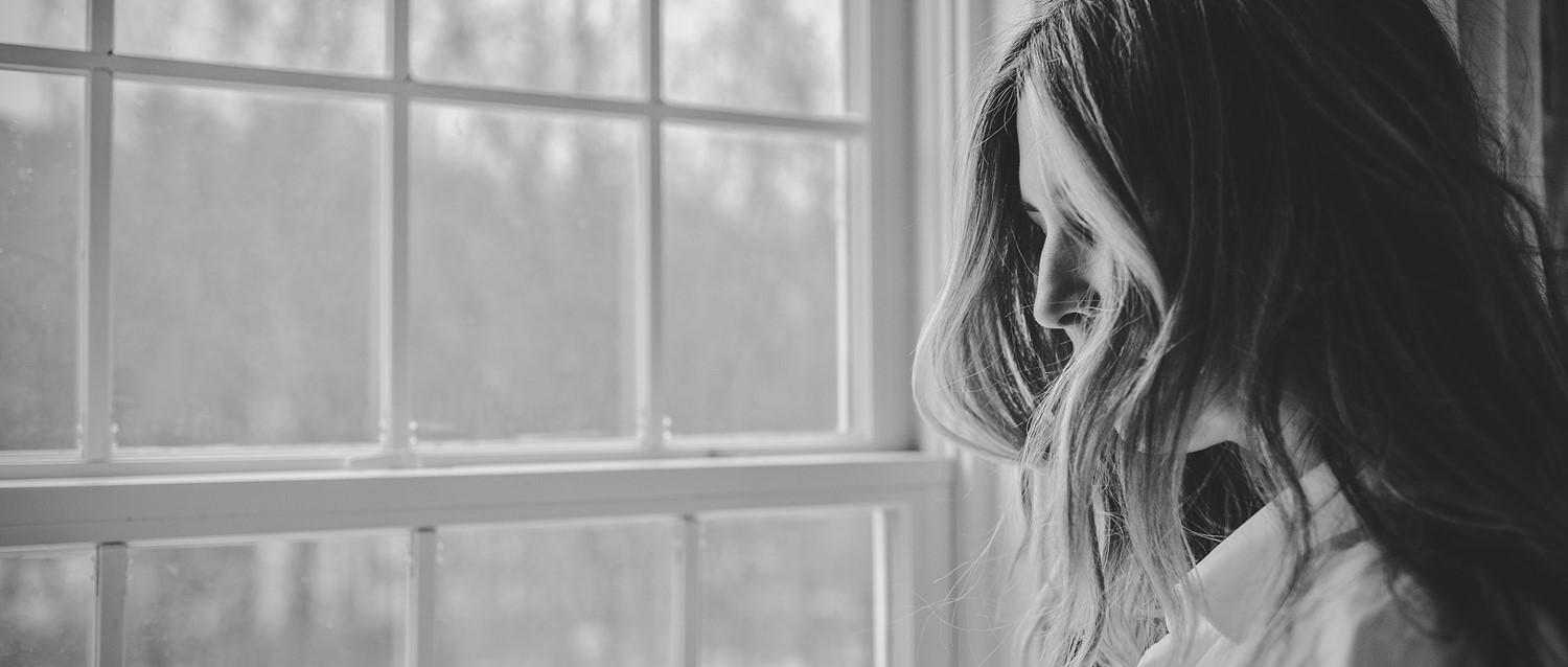 Personne aux cheveux longs de profil regardant vers le bas près d'une grande fenêtre, capturée en noir et blanc.