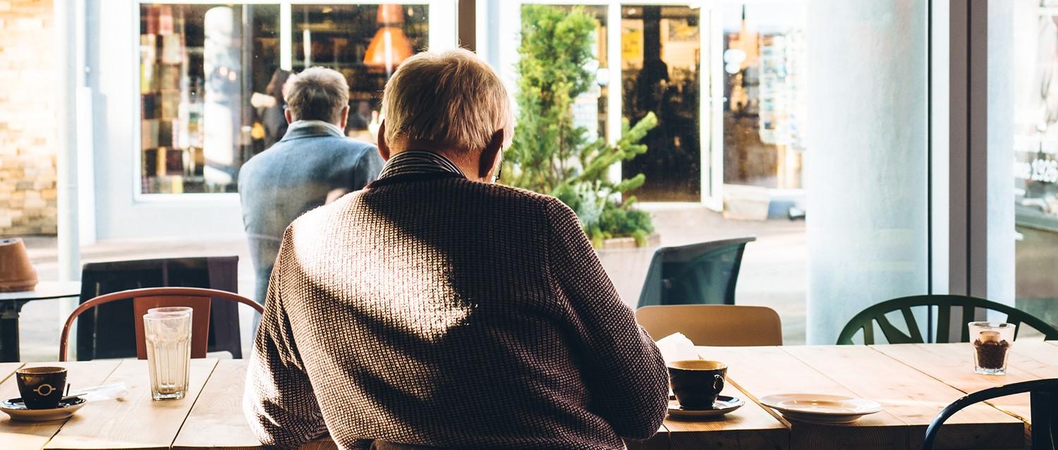 Deux personnes âgées assises dans un café lumineux, les tasses de café posées sur des tables en bois, regardant à travers de grandes fenêtres.