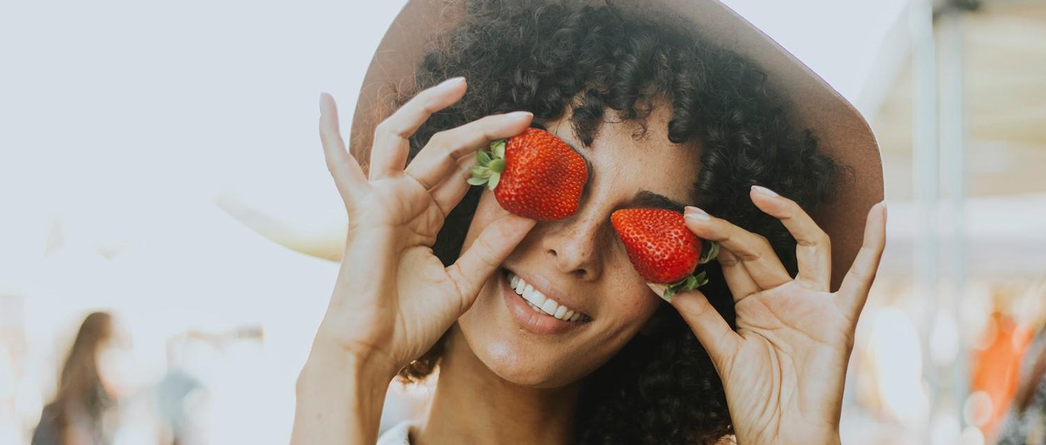 Personne aux cheveux bouclés tenant des fraises fraîches sur ses yeux, souriant de manière ludique dans une lumière naturelle douce.