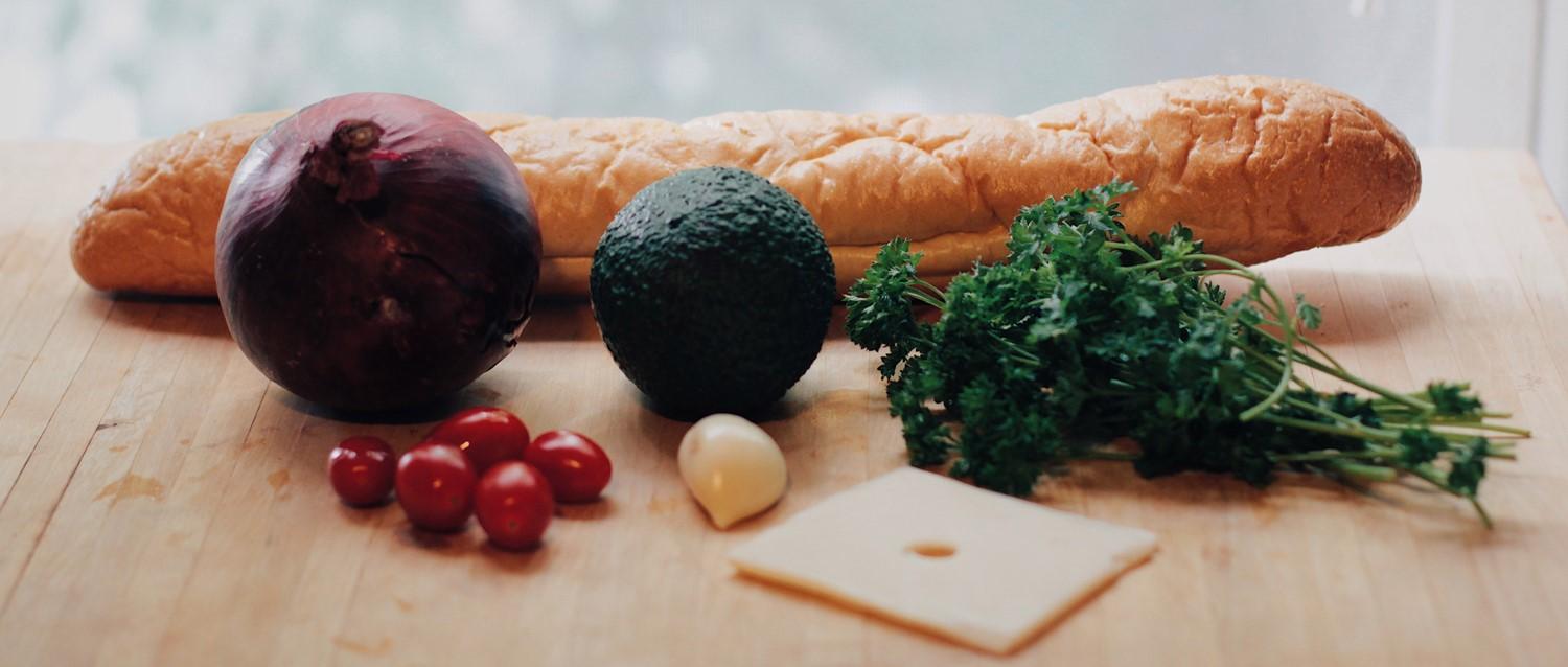 Ingrédients frais sur planche en bois : baguette, oignon rouge, avocat, persil, tomates cerises, ail et tranche de fromage.