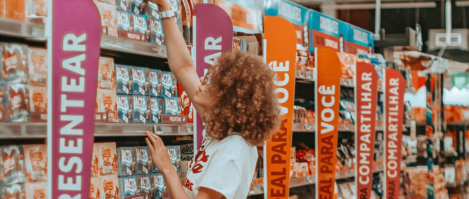 Personne aux cheveux bouclés cherchant des produits dans une allée de supermarché avec des panneaux de catégorie colorés en rose et orange.