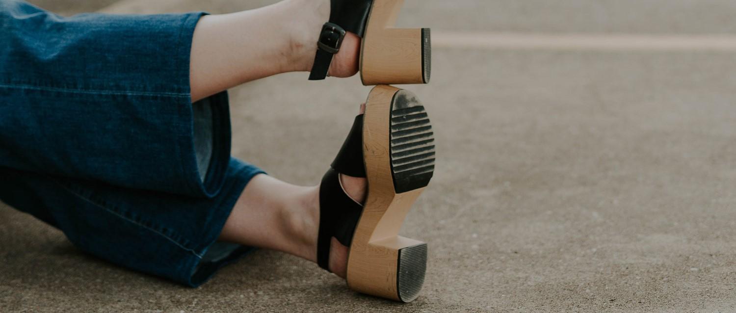 Personne portant un jean bleu et des sandales noires à talons en bois avec lipodème, avec une montre noire au poignet.