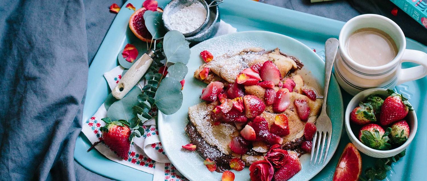 Plateau de petit-déjeuner avec des crêpes garnies de fraises fraîches, de sucre en poudre, de café et d'un petit bol de fruits.