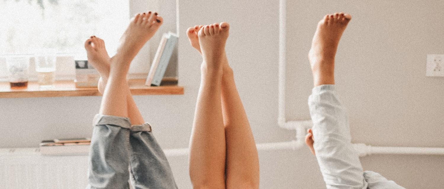 Trois paires de pieds nus levés en l'air contre le mur d'une salle de bain lumineuse, avec une baignoire visible au premier plan.