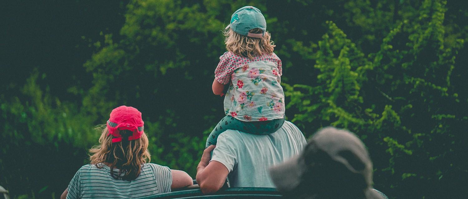 Child wearing floral shirt and teal cap sits on adult's shoulders, with another child in red cap nearby, against green trees.
